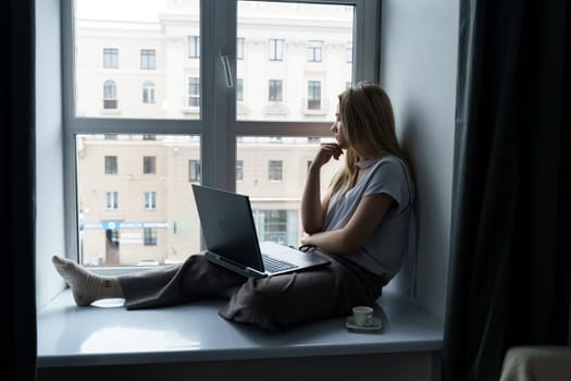 A young woman sits on a windowsill, works at a laptop, looks out the window. Business and education concept.