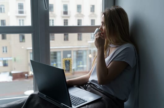 A young woman sits on a windowsill, drinks coffee, works at a laptop, looks out the window. Business and education concept. Close-up.
