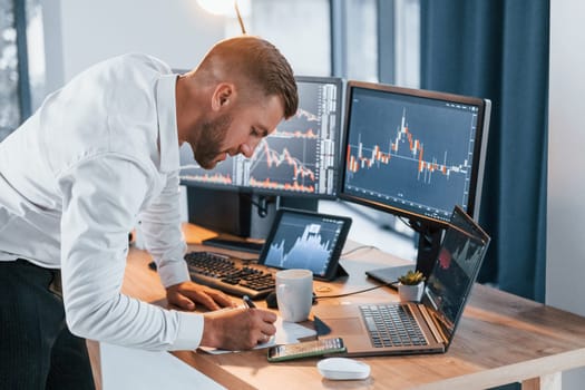 Near the table. Young businessman in formal clothes is in office with multiple screens.