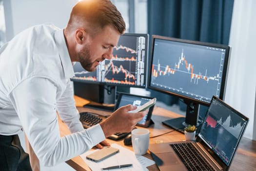 Near the table. Young businessman in formal clothes is in office with multiple screens.