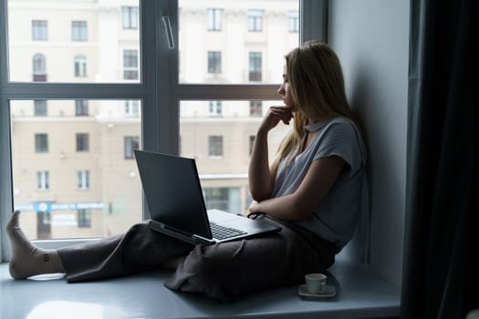 A young woman sits on a windowsill, works at a laptop, looks out the window. Business and education concept.