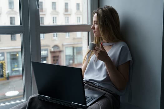 A young woman sits on a windowsill, drinks coffee, works at a laptop, looks out the window. Business and education concept. Close-up.