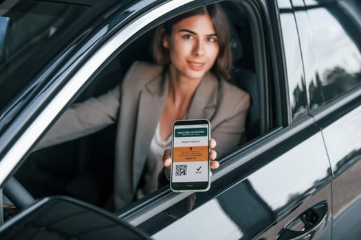 Holding vaccine passport. Woman testing new car. Sitting indoors in modern automobile.