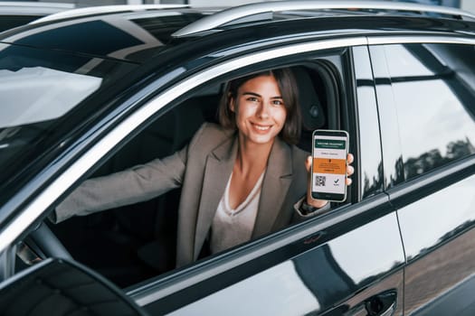 Holding smartphone with digital vaccine passport. Woman testing new car. Sitting indoors in modern automobile.
