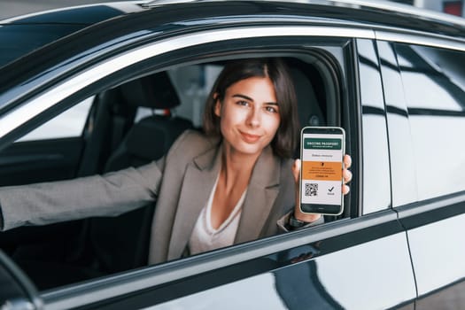 Holding smartphone with digital vaccine passport. Woman testing new car. Sitting indoors in modern automobile.