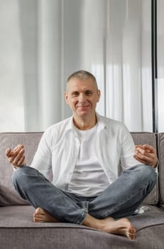 Portrait of a man who sits on a sofa in a room in a lotus position. The man is doing yoga.