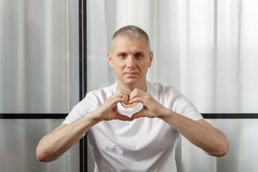 A middle-aged man in a white T-shirt smiles and shows a heart symbol.