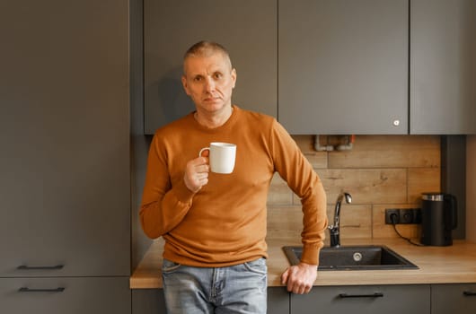 Portrait of a mature man in an orange jumper in the kitchen with a mug of warm tea.