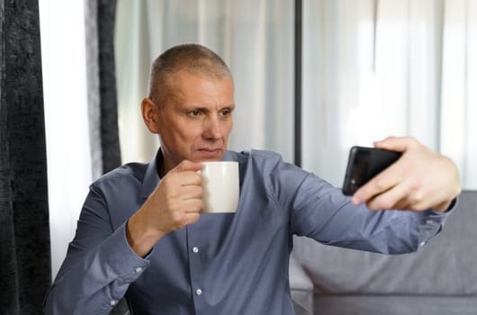 A middle-aged man with tea takes a selfie photo with a mobile phone in the apartment.
