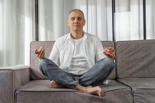 Portrait of a man who sits on a sofa in a room in a lotus position, meditates with his eyes closed. The man is doing yoga.