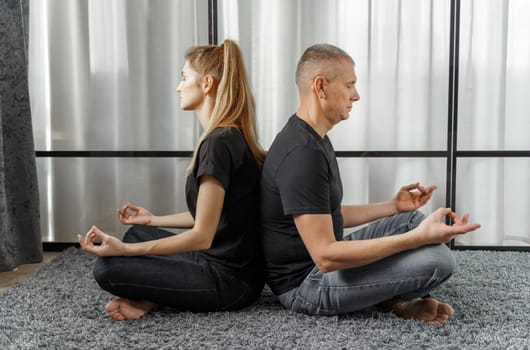 A man and a woman sit back to back, practice yoga and meditate for relaxation and balance of life in a room at home. Sports concept.