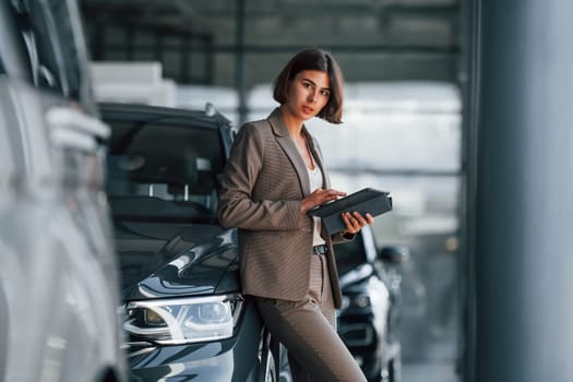 With tablet in hands. Woman is indoors near brand new automobile indoors.