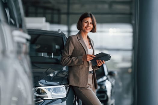 With tablet in hands. Woman is indoors near brand new automobile indoors.