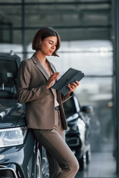 With tablet in hands. Woman is indoors near brand new automobile indoors.
