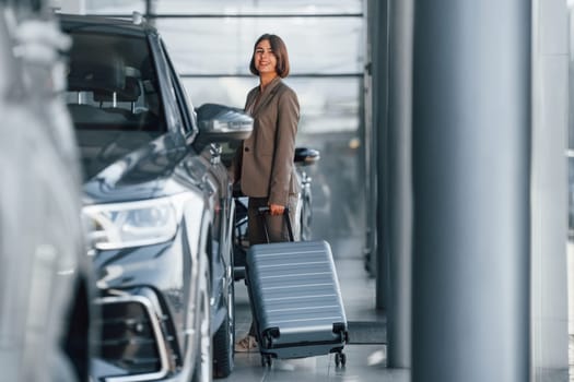 Holding luggage. Woman is indoors near brand new automobile indoors.