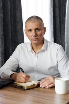 A middle-aged man sits at a table, works, writes down information in a notebook. Vertical frame. Remote work at home.