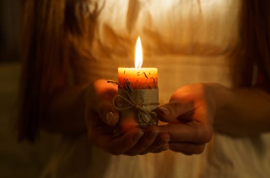 A woman holds a burning candle in her palms on a dark black background. Close-up of a burning candle.