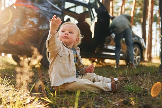 Girl is sitting on the ground. Happy family of father, mother and little daughter is in the forest.