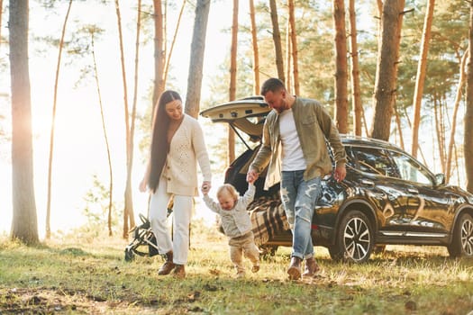 Against black automobile. Happy family of father, mother and little daughter is in the forest.