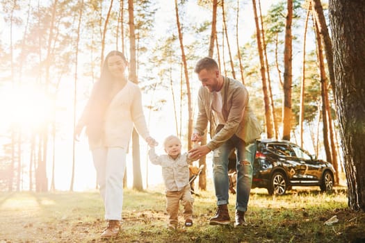 Against black automobile. Happy family of father, mother and little daughter is in the forest.