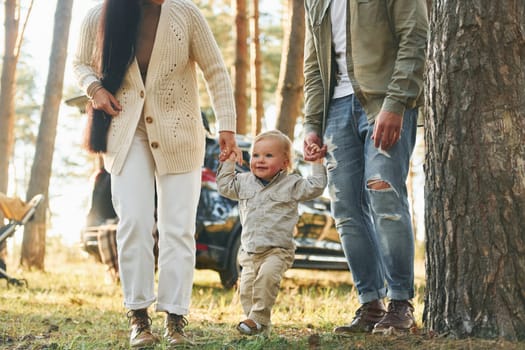 Against black automobile. Happy family of father, mother and little daughter is in the forest.