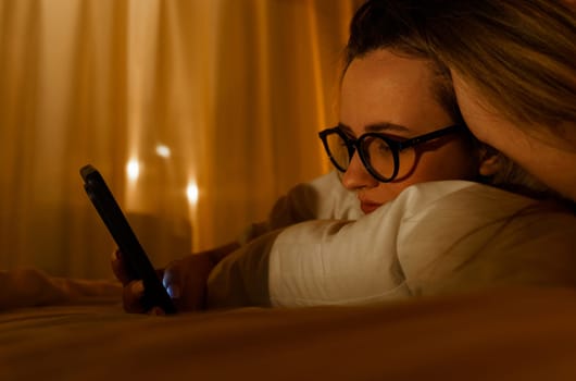 Young woman in glasses with smartphone lying in bed at home at night. Close-up