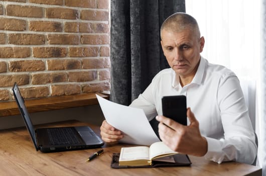 A businessman holds a phone and documents in his hands, reads information from the phone. Financial manager, company.