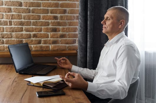 A businessman meditates while working on a laptop, sitting with his eyes closed at his workplace in a home interior. Work and rest.