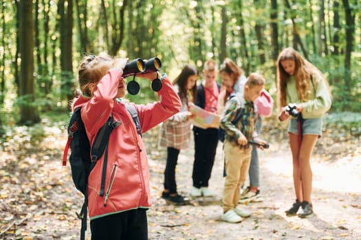 Girl looking into binoculars. Kids in green forest at summer daytime together.