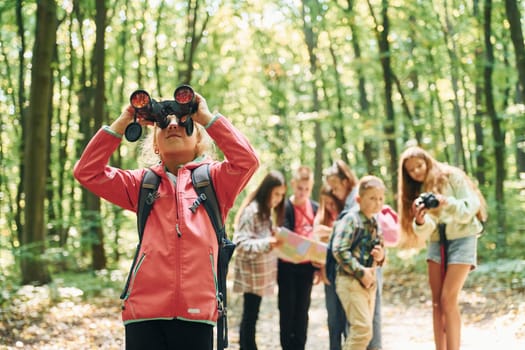 Girl looking into binoculars. Kids in green forest at summer daytime together.