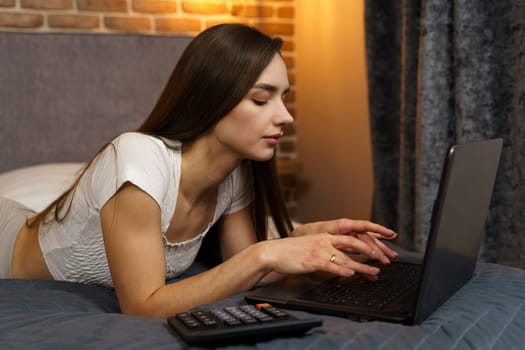 Happy beautiful woman lies on the bed and works on a laptop.