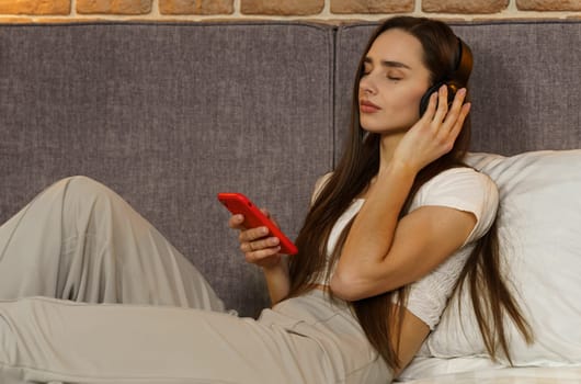 Happy young woman in headphones sits on the sofa and listens to music from a mobile phone, chooses tracks. Side view.