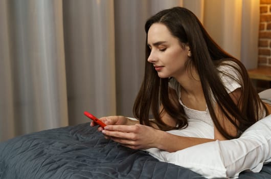 A young woman lies with a smartphone in her hands in the evening on the bed. Watching videos, social networks, reading messages.