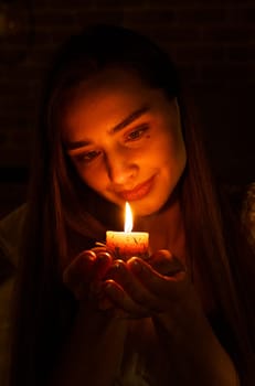 A woman holds a burning candle in her palms on a dark black background.