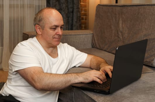 A mature man sits on the floor and works on a laptop that is on the sofa.