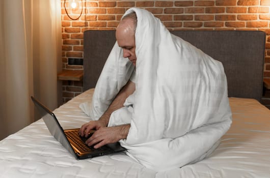 A man is sitting on a bed covered with a blanket, working at a computer.