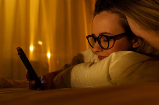Young woman in glasses with smartphone lying in bed at home at night. Close-up