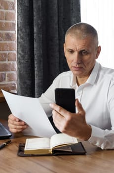 A businessman holds a phone and documents in his hands, reads information from the phone. Financial manager, company.