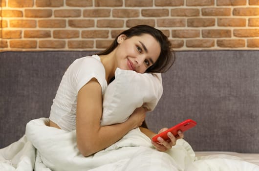 A young beautiful woman sits on a bed hugging a pillow, holding a phone in her hands, looking at the camera.