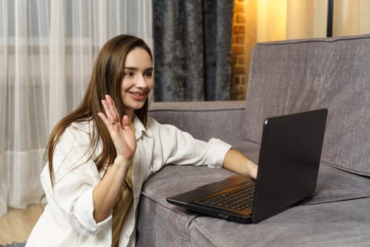 A beautiful young woman is talking on a video link on a laptop that is on the sofa, she is sitting on the floor herself.