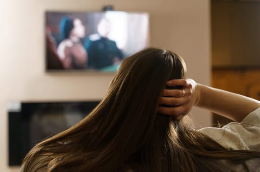The girl watches TV, sits in the living room at home in the evening, switches TV channels. View over the shoulder, the image is blurry.