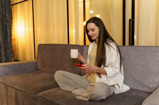 Young woman chatting with friends, surfing on the phone, enjoying her weekend at home with a cup of tea.