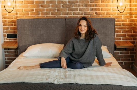 An attractive woman is sitting on a bed in a bedroom wearing blue trousers and a gray jumper. Smiling and looking at the camera.