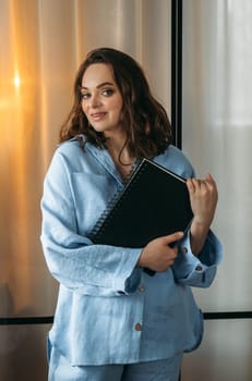 A portrait of a young woman in a light blue suit is standing in a room with a tablet and a notebook in her hands. Looks into the camera. Vertical frame.