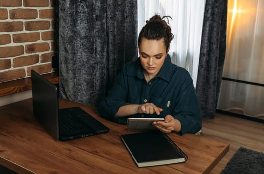 A beautiful woman sits at a table indoors and works remotely on a tablet.