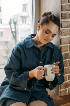 Portrait of a beautiful woman who is sitting on the windowsill indoors with a white cup in her hands. Vertical frame.