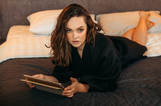 Portrait of a beautiful woman in a black bathrobe, who lies in bed with a tablet in her hands. Looks into the camera.
