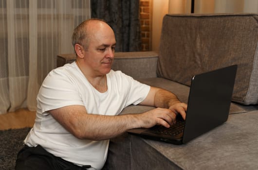 A mature man sits on the floor and works on a laptop that is on the sofa.
