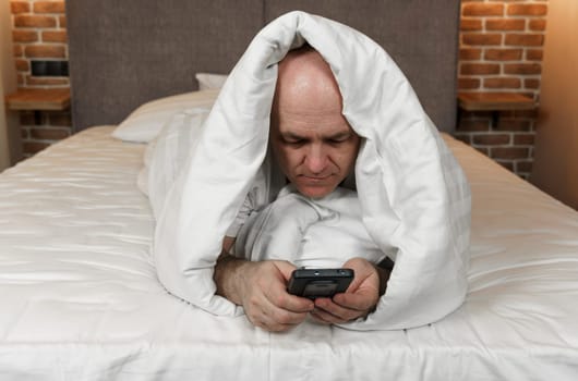 An adult man lies covered with a blanket and browses social networks, videos in a smartphone.