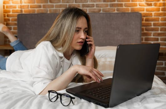 A young beautiful woman is lying on the bed, talking on the phone, looking into a laptop.